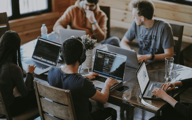 5 people sat around a table using laptops and talking to each other.