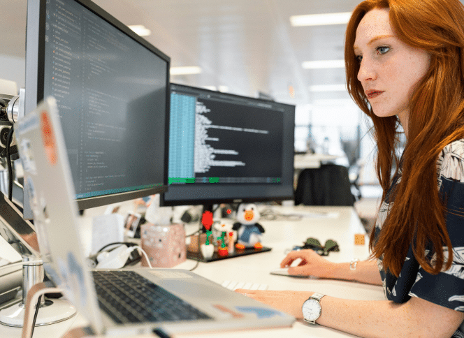 A woman sat at her desk with two computer screens, coding.