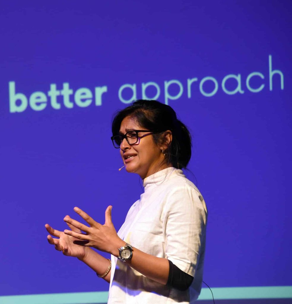 A woman speaking on stage with a blue wall behind her that says better approach.