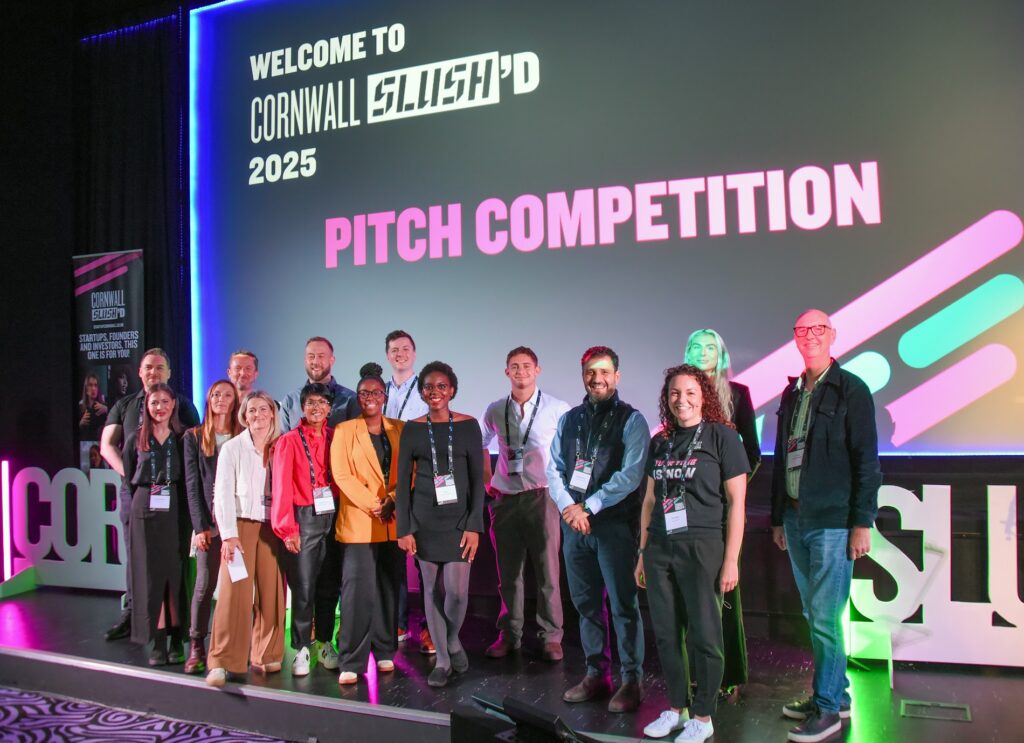 Image shows 15 people stood on a stage for the pitch competition at Cornwall Slush'd. There are 9 pitch finalists, the host of the day and the judges of the competition. Everyone is smiling at the camera.